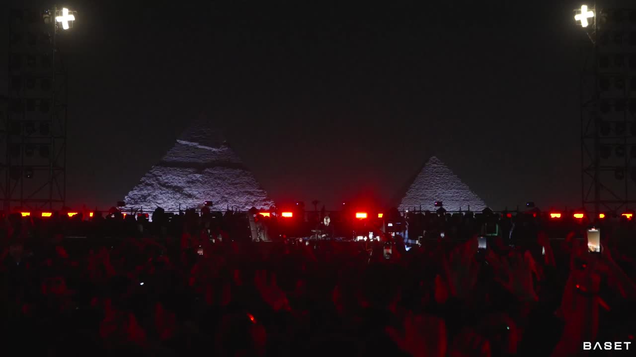 A vast festival crowd beneath the pyramids of Giza, washed in red light